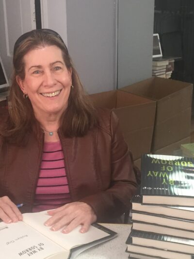 Robyn Gigl smiles for the camera as she sits behind a table of stacked books to her left, and has a book open to sign it.