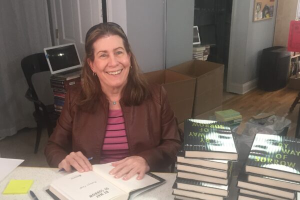 Robyn Gigl smiles for the camera as she sits behind a table of stacked books to her left, and has a book open to sign it.
