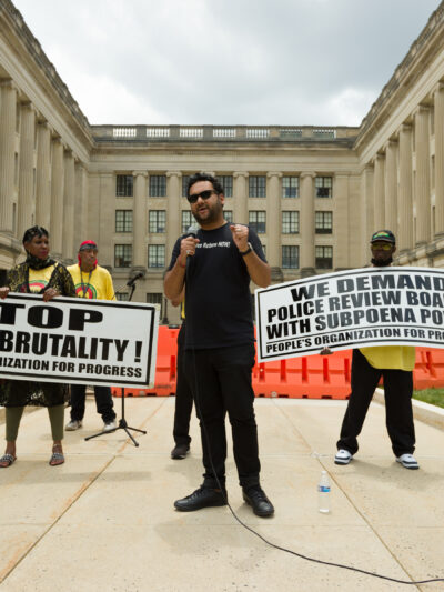 Amol SInha is speaking into a microphone at the State House. Two people are standing behind him holding signs that read "stop police brutality" and "we demand  police review boards with subpoena power"