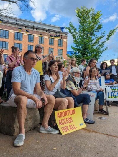 A crowd of people are gathered at a park, clapping and holding signs that read "Abortion Access for All"