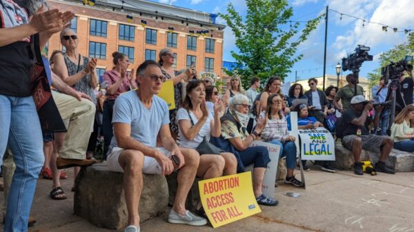 A crowd of people are gathered at a park, clapping and holding signs that read "Abortion Access for All"