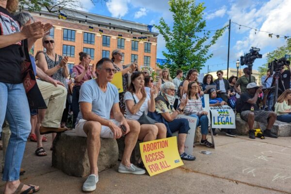 A crowd of people are gathered at a park, clapping and holding signs that read "Abortion Access for All"