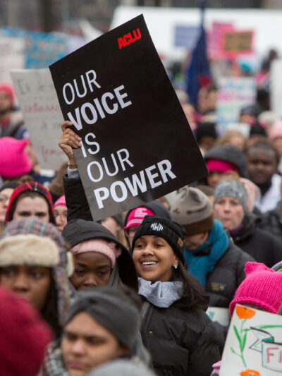 A smiling woman is holding a sign that says "Our voice is our power" in a crowded protest.