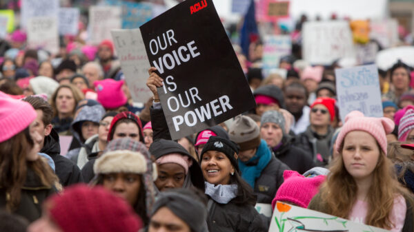 A smiling woman is holding a sign that says "Our voice is our power" in a crowded protest.