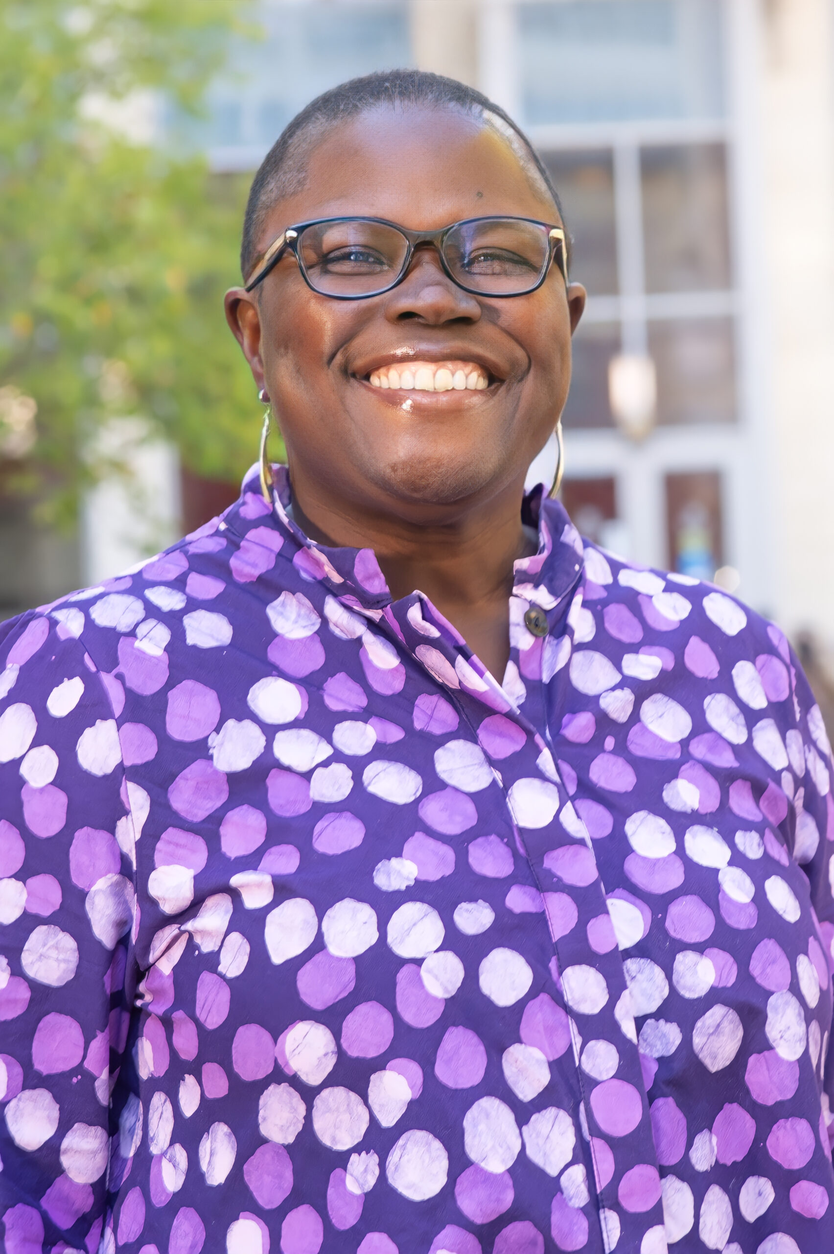 Kim Mutcherson, a black woman with shaved hair and glasses wearing a purple and white polka dot shirt, poses and smiles for a headshot image outdoors.