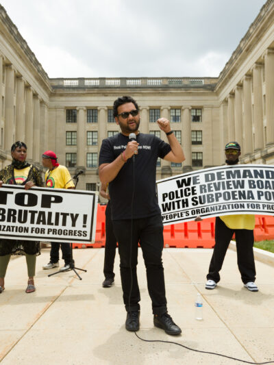 Amol SInha is speaking into a microphone at the State House. Two people are standing behind him holding signs that read "stop police brutality" and "we demand  police review boards with subpoena power"