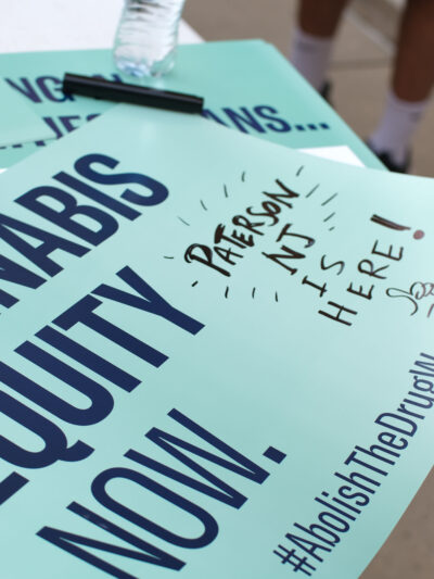 A mint green sign reads: Cannabis equity now. Hashtag Abolish the Drug War. A handwritten note is written off the side that reads: Paterson NJ is here!