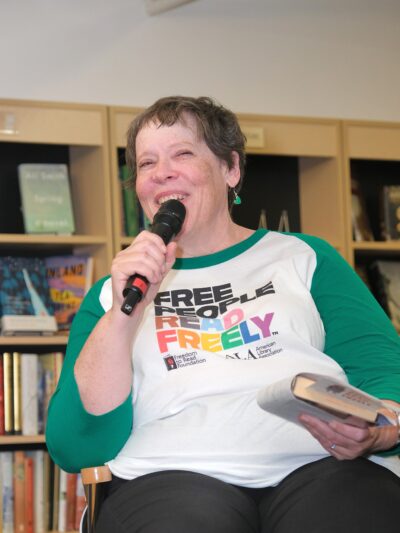 white Woman in a white and green tshirt holding a mircophone and a book addressing a crowd in a bookstore