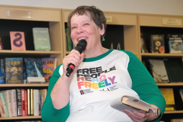 white Woman in a white and green tshirt holding a mircophone and a book addressing a crowd in a bookstore