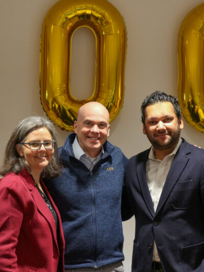 Three people standing in front of ballons of the number 100 in an office conference room