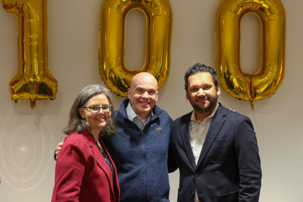 Three people standing in front of ballons of the number 100 in an office conference room