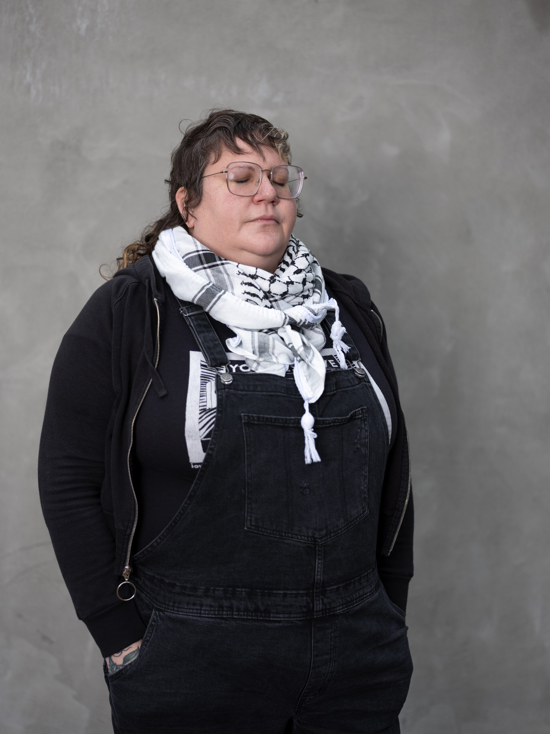 white non-binary person wearing a black shirt and a black/white scarf standing with their hands in their pockets in front of a grey background