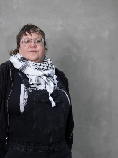 White woman wearing a black shirt and a scarf standing in front of a grey background