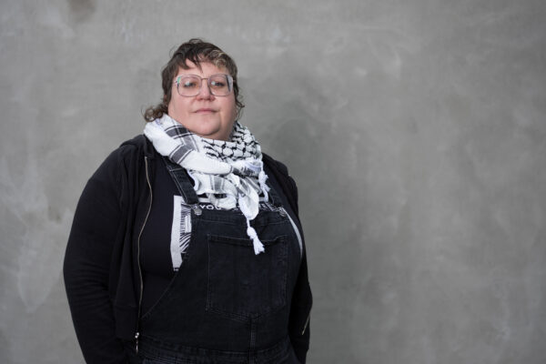 White woman wearing a black shirt and a scarf standing in front of a grey background