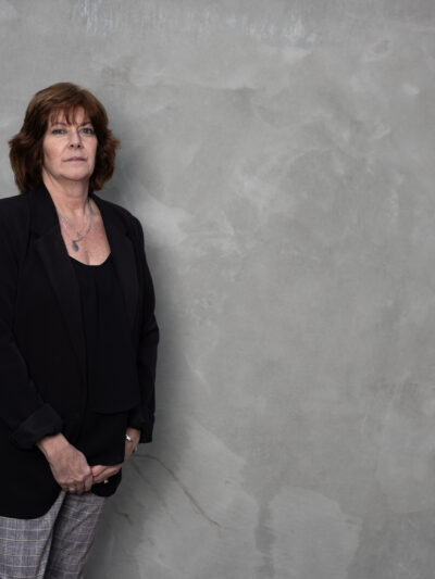 White women wearing a black blazer and grey pants in front of a grey background
