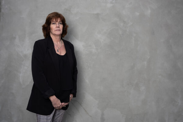 White women wearing a black blazer and grey pants in front of a grey background