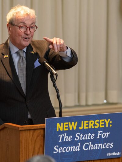 Governor Phil Murphy speaking at a podium with a sign that reads "New Jersey: The State for Second Chances"