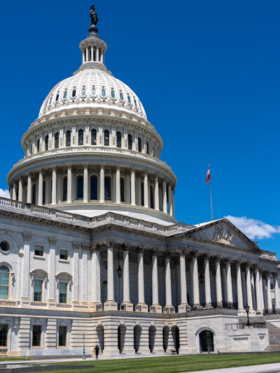 Wide daytime view of the U.S. Capitol building in Washington, D.C., with the white dome and columns under a bright blue sky.
