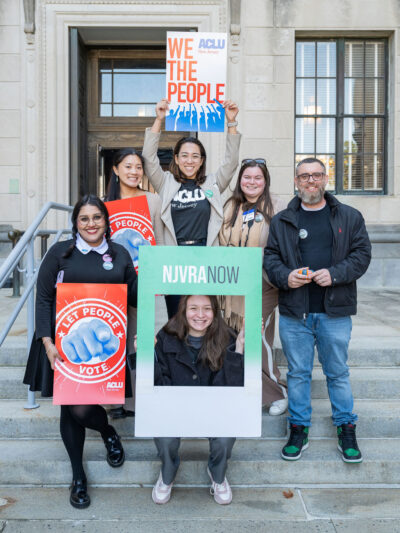 People stand on stairs holding signs that say "We the People" "NJVRA Now" and "Let People Vote"