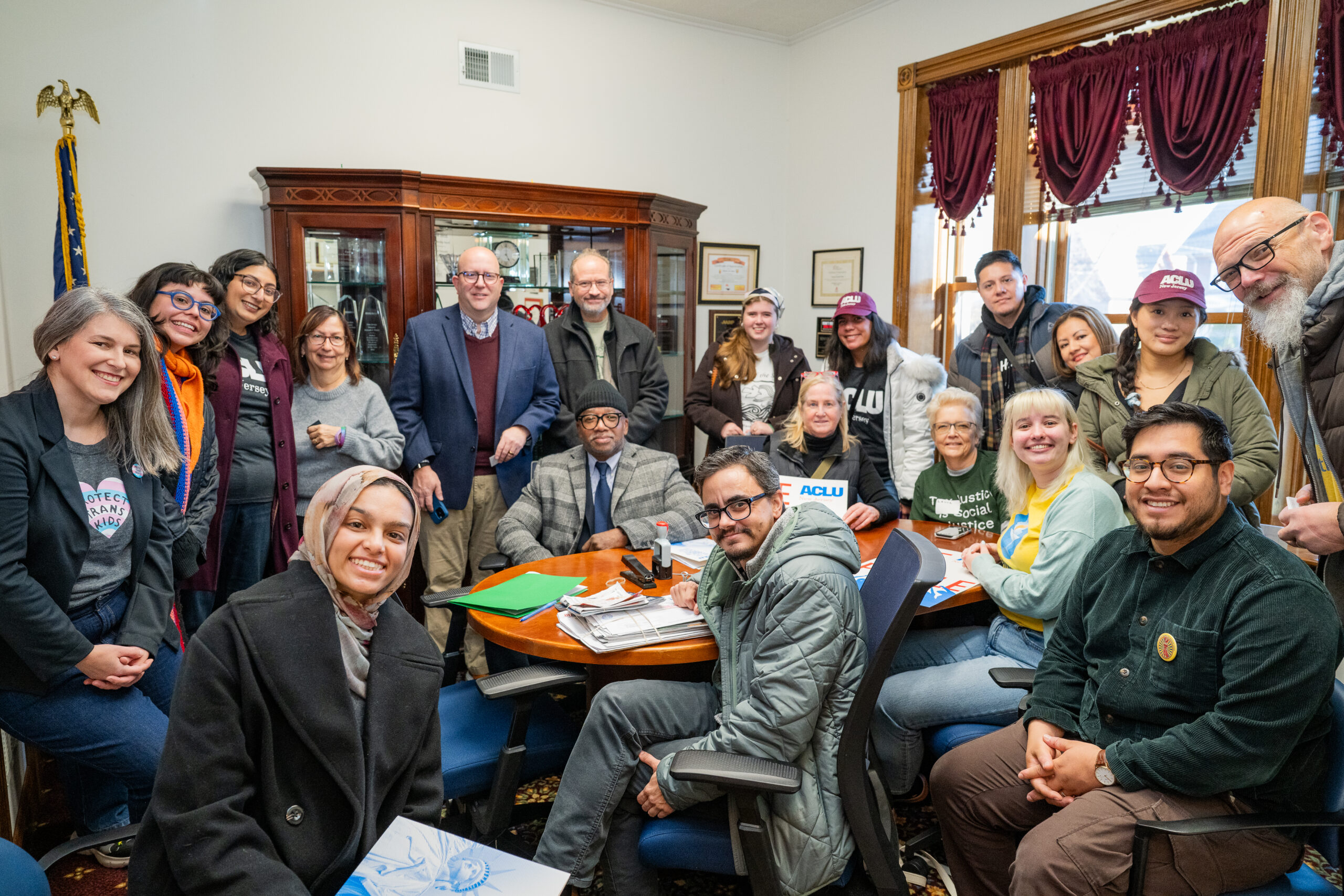Advocates participate in an in-district lobby day in support of the People’s Agenda on December 2, 2025. A group of these advocates are gathered in an office, smiling at the camera. Some are seated at a table, while others stand around them. The room has a U.S. flag and a cabinet with glass doors, and there are ACLU signs visible.