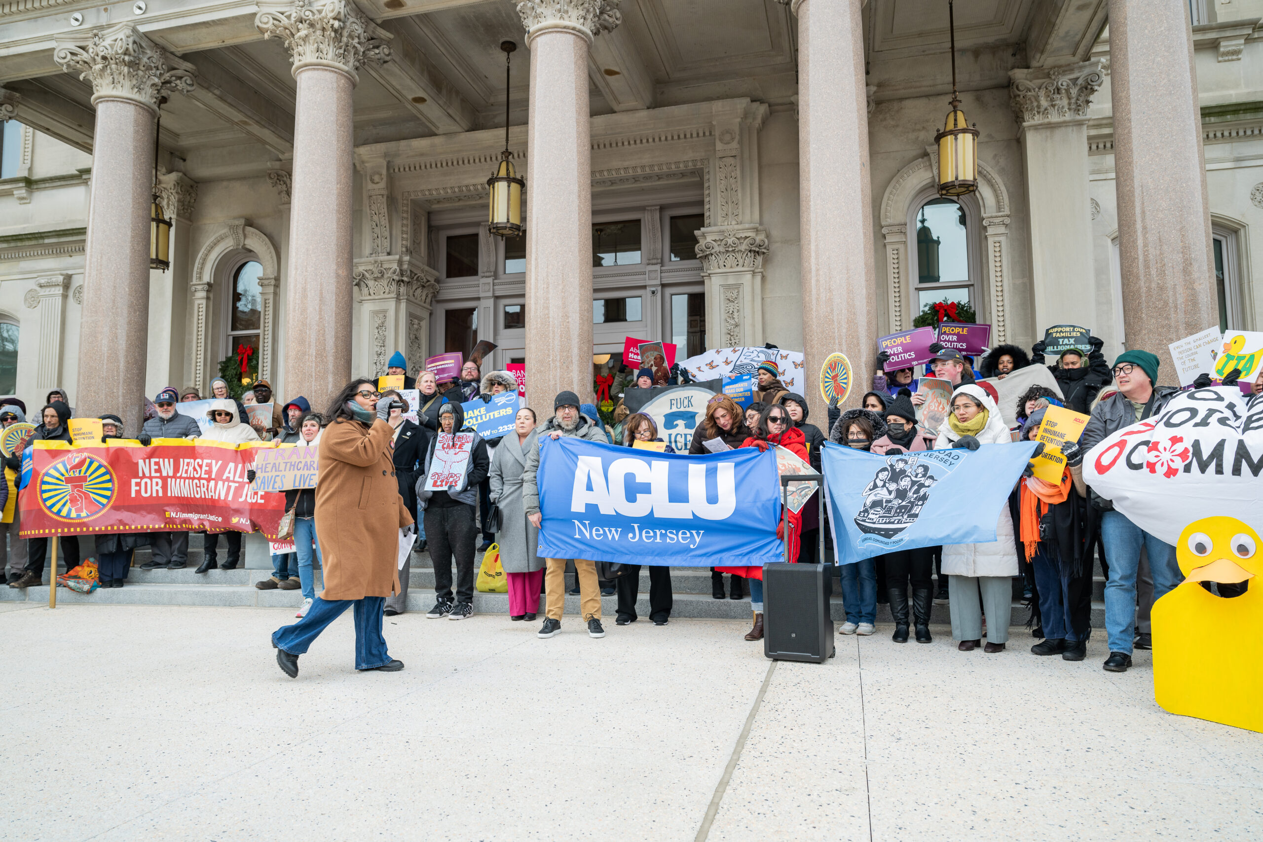 Advocates rally outside of the New Jersey Statehouse in support of the People’s Agenda on December 8, 2025. A group of people in winter clothing stand on the steps of a building holding signs and banners, including one from the ACLU-NJ.