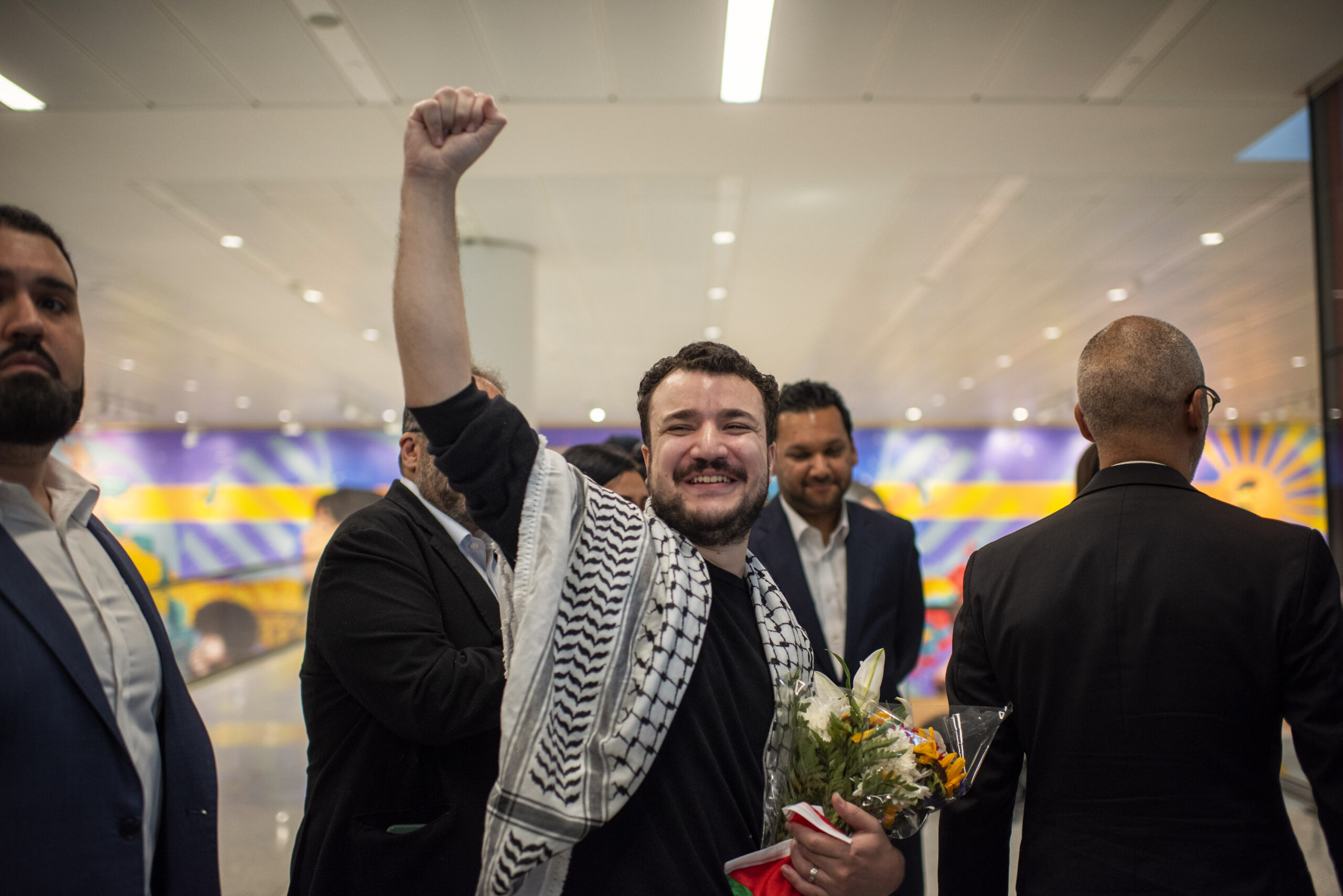 After 104 days of being unjustly detained for his advocacy for Palestinian rights, Mahmoud Khalil arrives home. He is shown smiling and raising a fist in celebration, surrounded by a group of people at the airport while holding a bouquet of flowers.