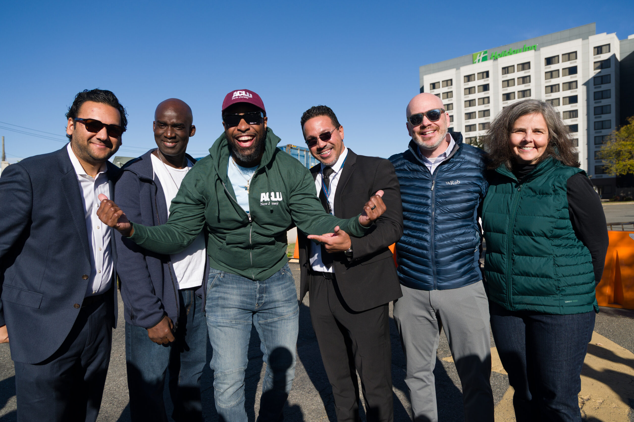 Jamse Comer celebrates his release with friends, family, and the ACLU-NJ on October 17,2025. James stands posing and smiling with a group of people on a sunny day wearing a maroon baseball cap and dark green hoodie with the ACLU-NJ logo.
