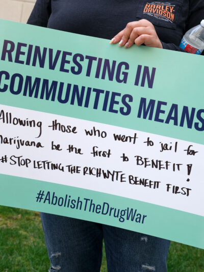 A person holds a sign reading "Reinvesting in Communities Means... Allowing those who went to jail for marijuana be the first to benefit!! #StopLettingTheRichWyteBenefitFirst #AbolishTheDrugWar." Below, text states "New Jersey must live up to its promise of equitable cannabis reform." The image features the ACLU NJ logo.