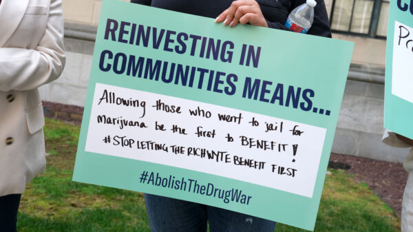 A person holds a sign reading "Reinvesting in Communities Means... Allowing those who went to jail for marijuana be the first to benefit!! #StopLettingTheRichWyteBenefitFirst #AbolishTheDrugWar." Below, text states "New Jersey must live up to its promise of equitable cannabis reform." The image features the ACLU NJ logo.