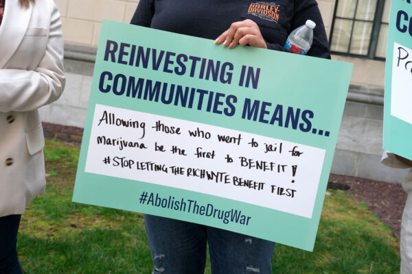 A person holds a sign reading "Reinvesting in Communities Means... Allowing those who went to jail for marijuana be the first to benefit!! #StopLettingTheRichWyteBenefitFirst #AbolishTheDrugWar." Below, text states "New Jersey must live up to its promise of equitable cannabis reform." The image features the ACLU NJ logo.
