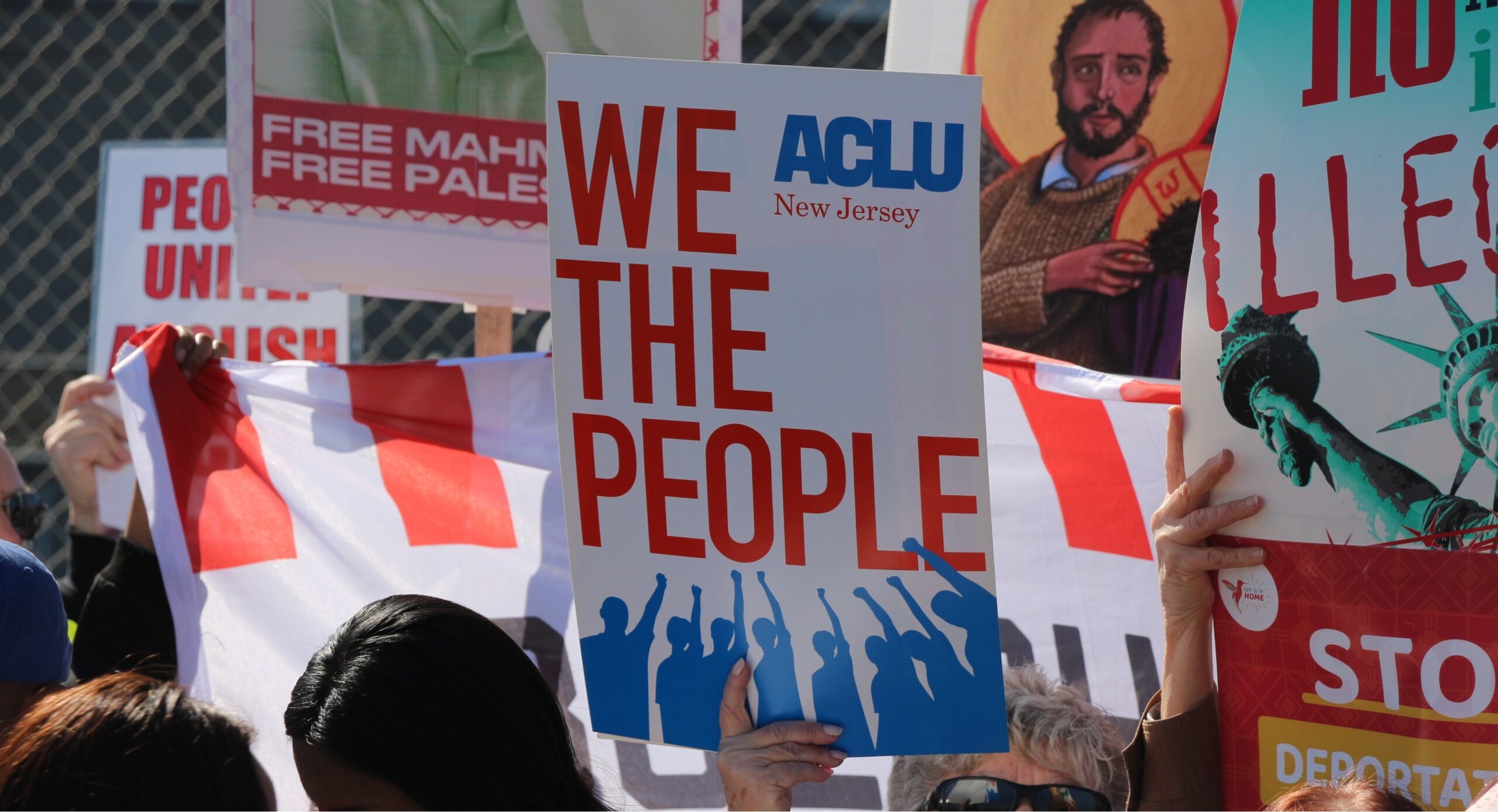 A group of people holding protest signs. The prominent sign says "We the People" with the ACLU New Jersey logo.