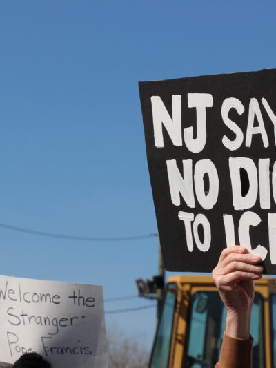 Two protest signs are held up against a clear blue sky. One sign reads, "NJ SAYS NO DICE TO ICE!" The other sign says, "Welcome the Stranger - Pope Francis." A construction vehicle and utility wires are visible in the background.