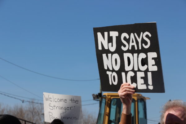 Two protest signs are held up against a clear blue sky. One sign reads, "NJ SAYS NO DICE TO ICE!" The other sign says, "Welcome the Stranger - Pope Francis." A construction vehicle and utility wires are visible in the background.