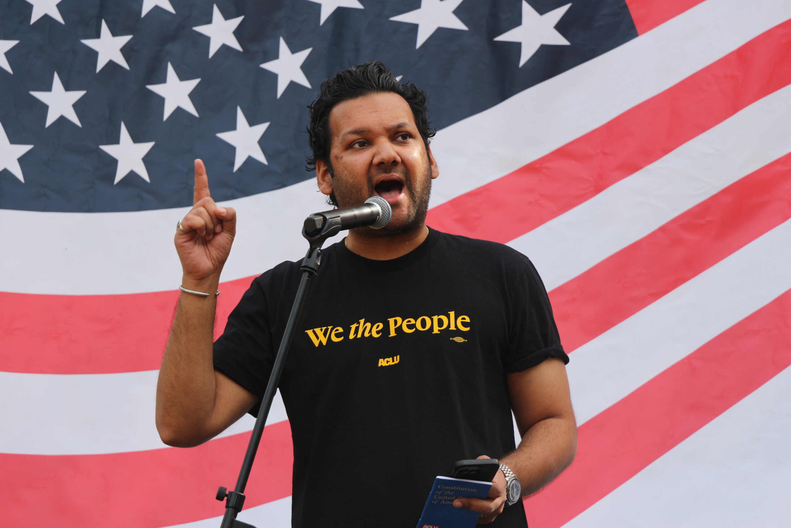 ACLU-NJ Executive Director Amol Sinha speaks passionately into a microphone at a No Kings rally on October 18,2025. He is wearing a "We the People" shirt in front of a large American flag backdrop.