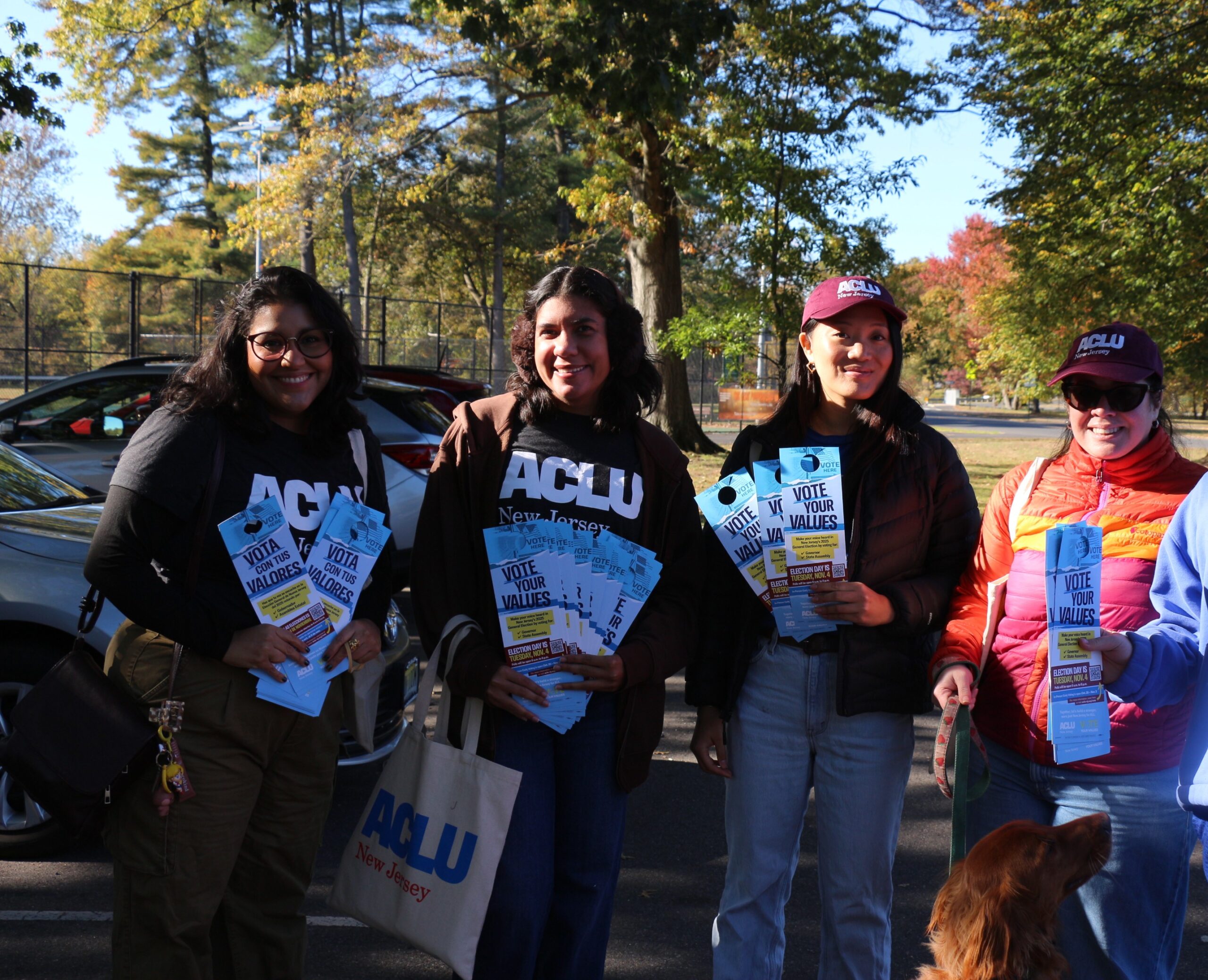 A group of four people is standing outside holding flyers and an ACLU-NJ tote bag. They are preparing to canvass a neighborhood in New Jersey. A few of them are wearing ACLU-NJ apparel. Trees and cars are visible in the background