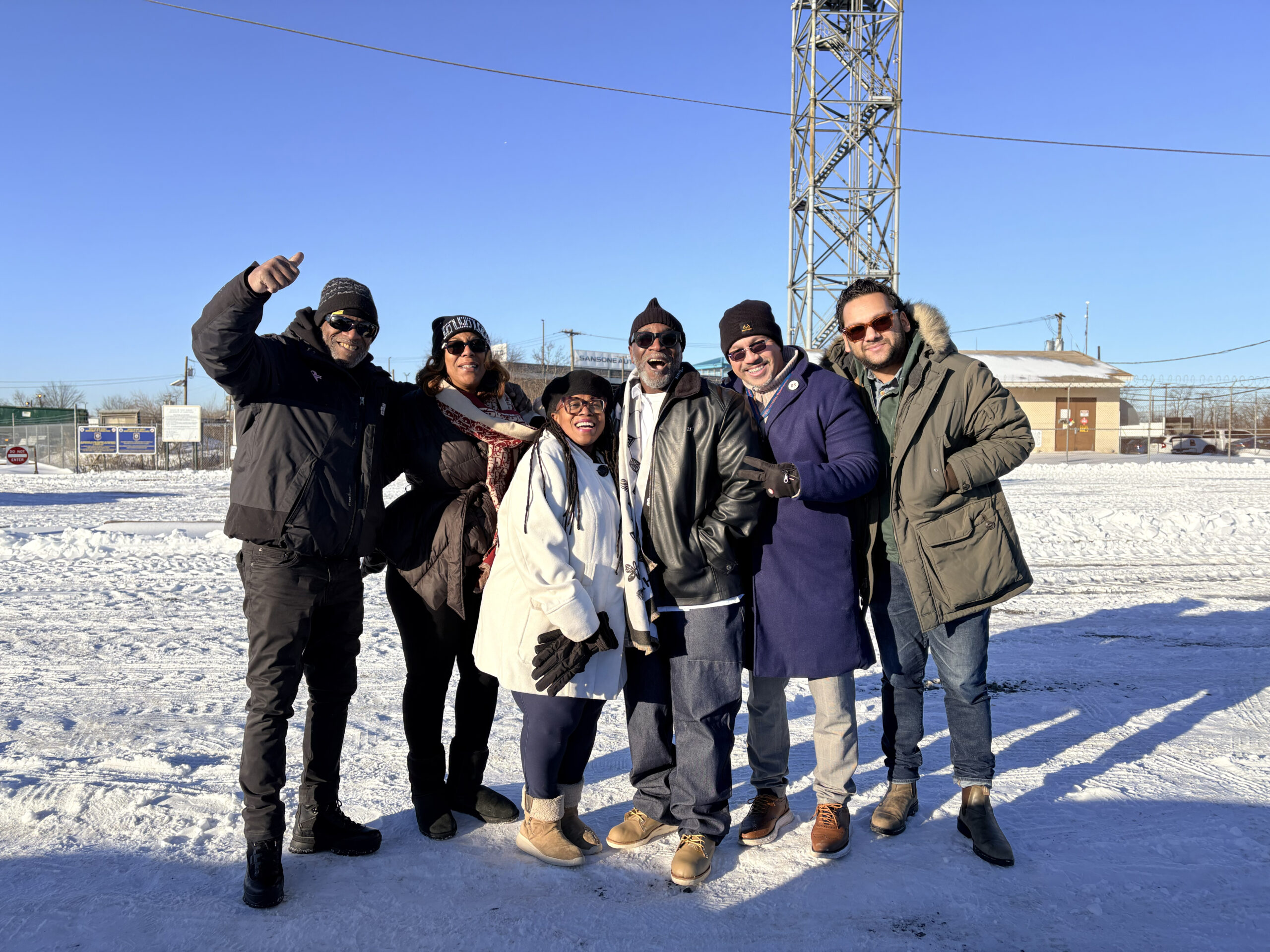 ACLU-NJ greeted Clemency Project clients released from East jersey State Prison on January 30, 2026. A group of six people dressed in winter clothing stand together on a snowy ground with clear blue skies above. A metal tower and some buildings are visible in the background.