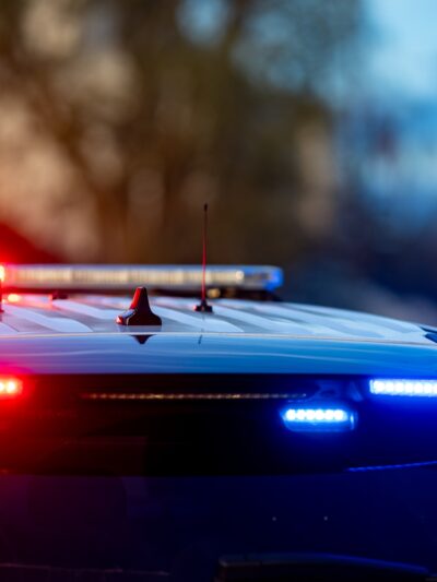 A police car with flashing red and blue lights is parked on a street. The background shows a blurred view of trees and a building at dusk.