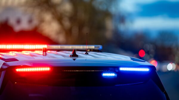 A police car with flashing red and blue lights is parked on a street. The background shows a blurred view of trees and a building at dusk.