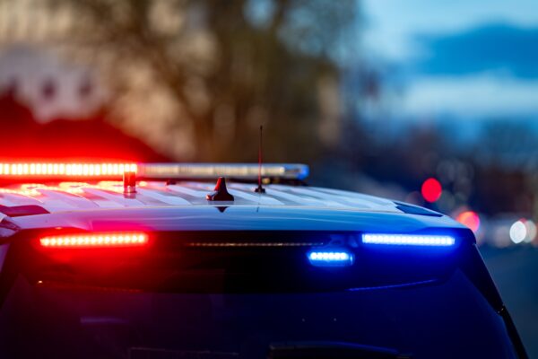A police car with flashing red and blue lights is parked on a street. The background shows a blurred view of trees and a building at dusk.