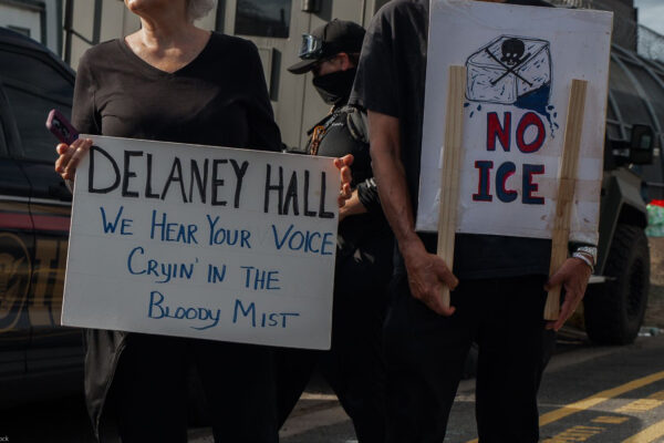 People hold signs outside the Delaney Hall Detention Facility during a protest against the treatment of detainees; one sign reads "Delaney Hall, we hear your voice cryin’ in the bloody mist," and another says "NO ICE" beneath a skull-and-crossbones drawing, with a masked officer and parked vehicles behind them.