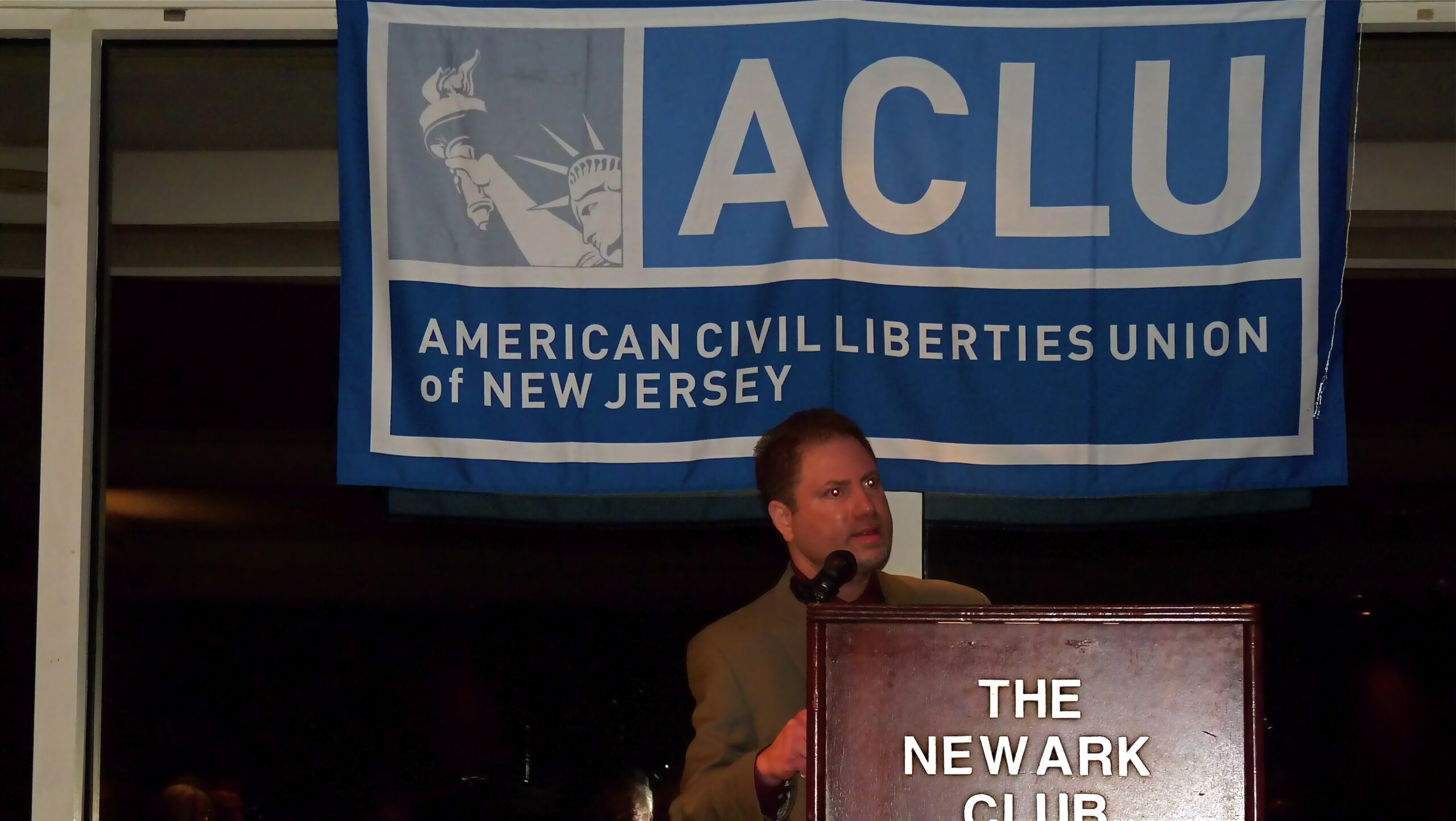 A man giving a speech in front of a banner with the ACLU-NJ logo on it.