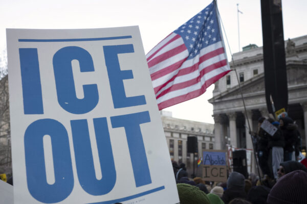 A hand holds a large white protest sign reading "ICE OUT" in bold blue letters during a crowded demonstration outside a government building. An American flag waves above the crowd, and additional signs in the background include messages calling to abolish ICE.