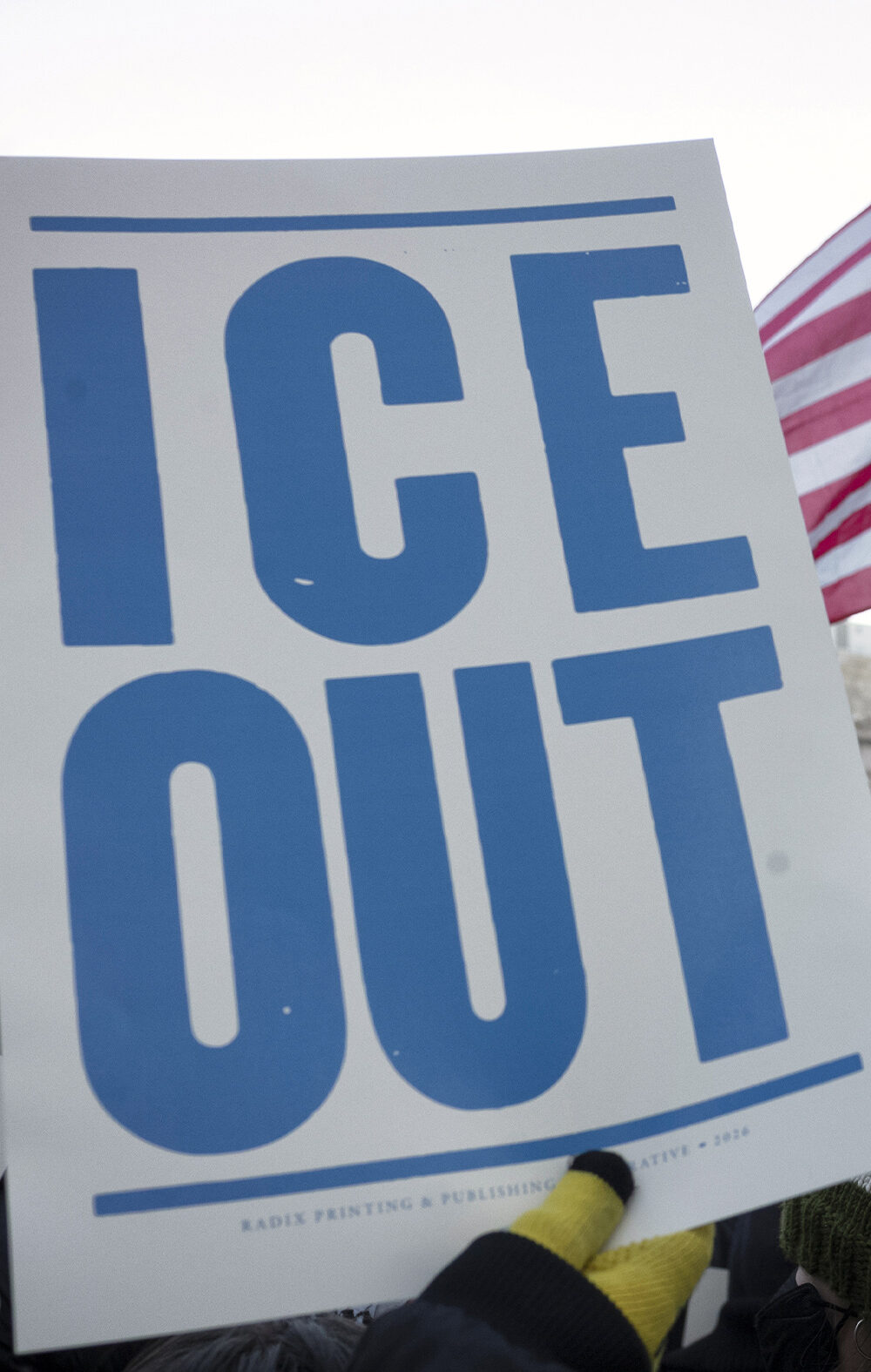A hand holds a large white protest sign reading "ICE OUT" in bold blue letters during a crowded demonstration outside a government building. An American flag waves above the crowd, and additional signs in the background include messages calling to abolish ICE.