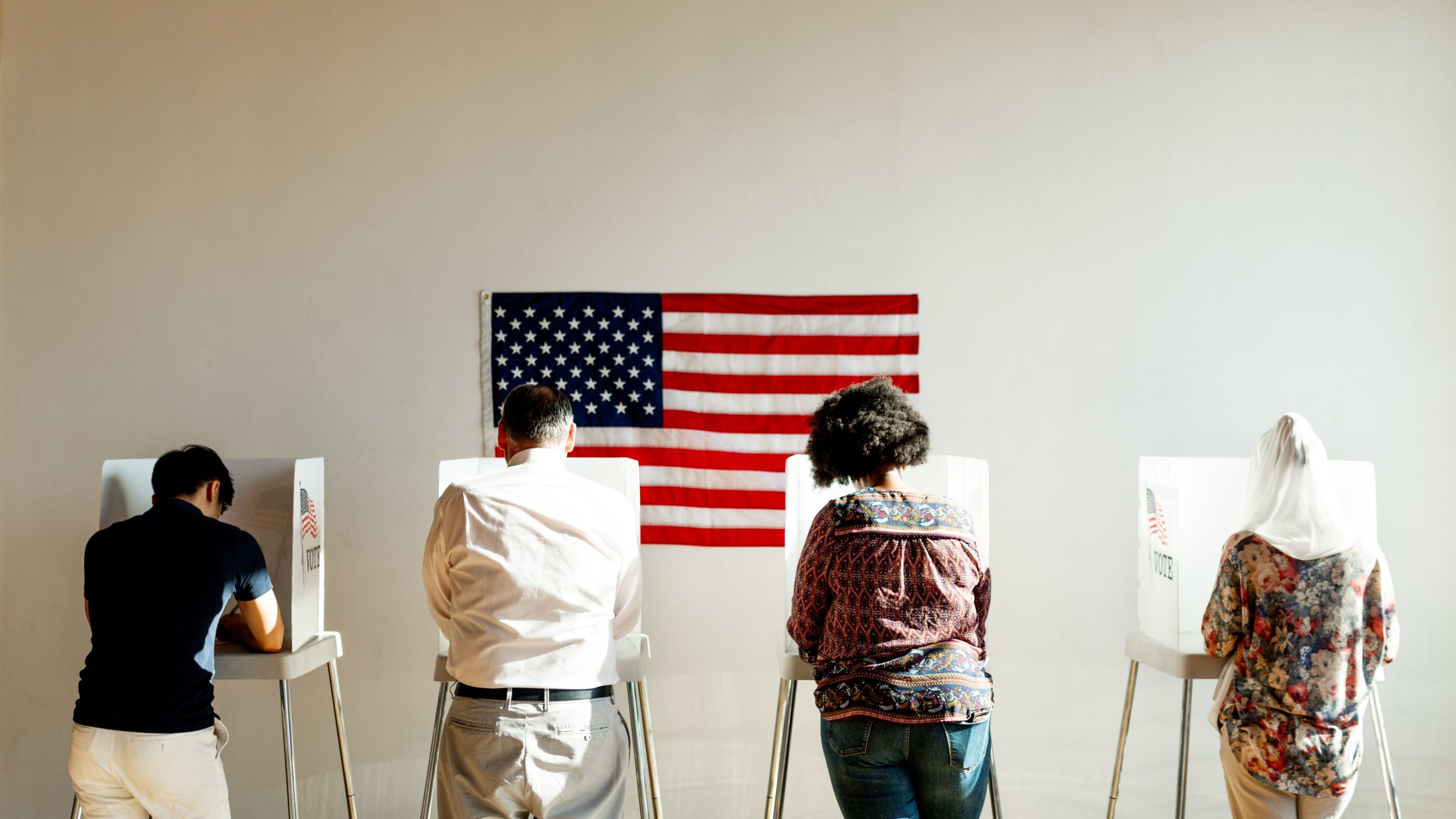 A group of people voting in polling booths with an American Flag behind them