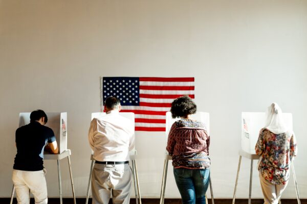 A group of people voting in polling booths with an American Flag behind them