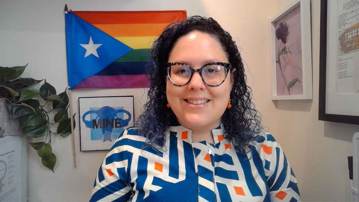 Dr. Kristyn Brandi, a white woman with medium length curly hair and glasses, is smiling in an office setting, with art and plants in the background