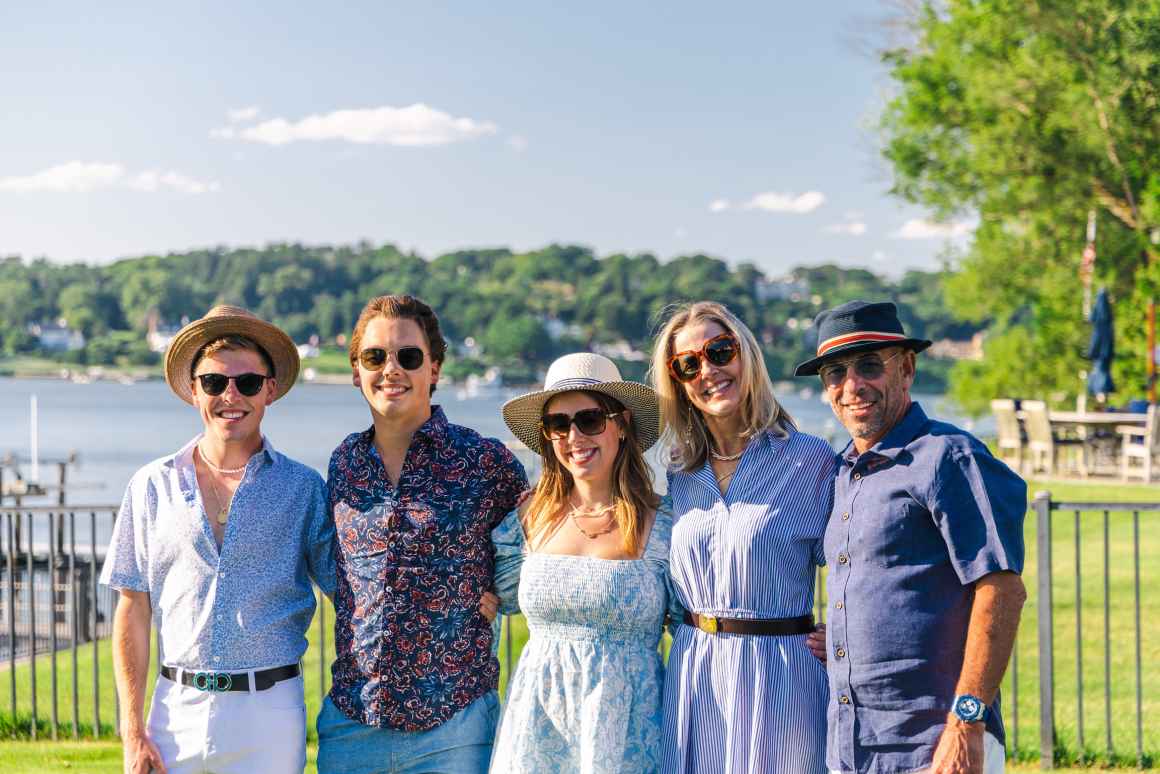A family of five dressed in blue and sunglasses is smiling at the camera. In the background, there is a body of water and greenery.
