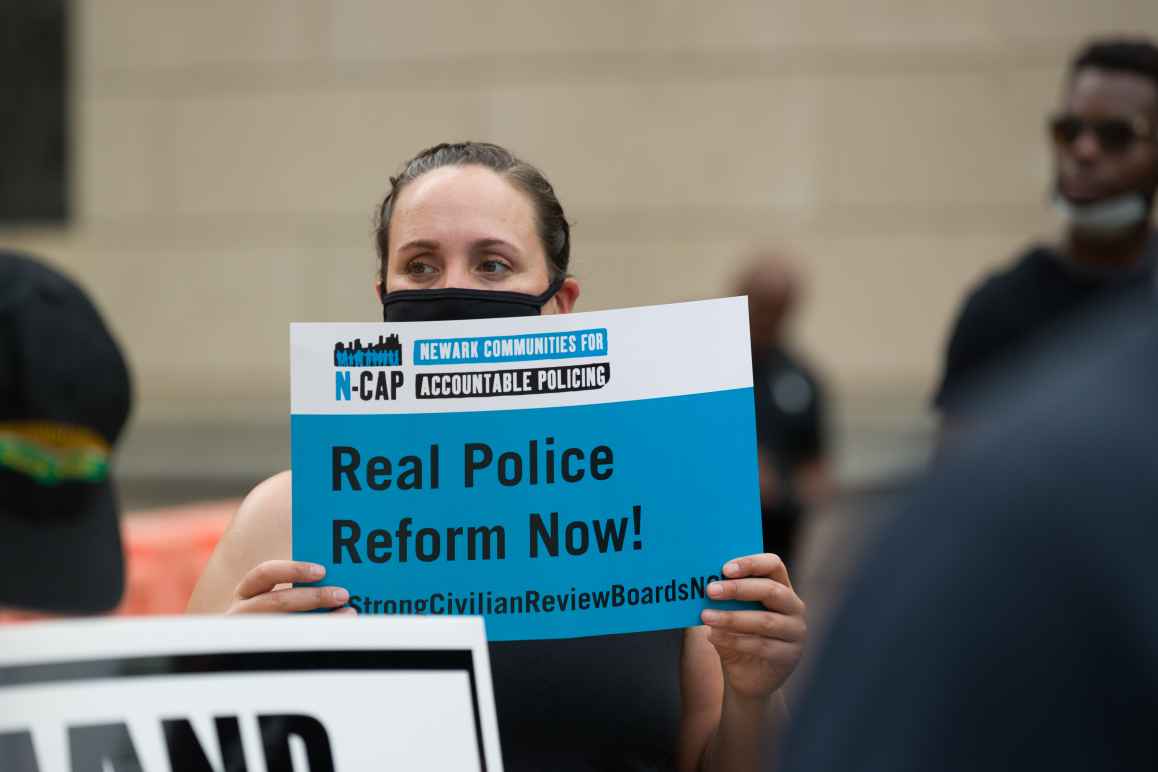 A masked woman holds a sign by Newark Communities for Accountable Policing that reads: Real Police Reform Now!
