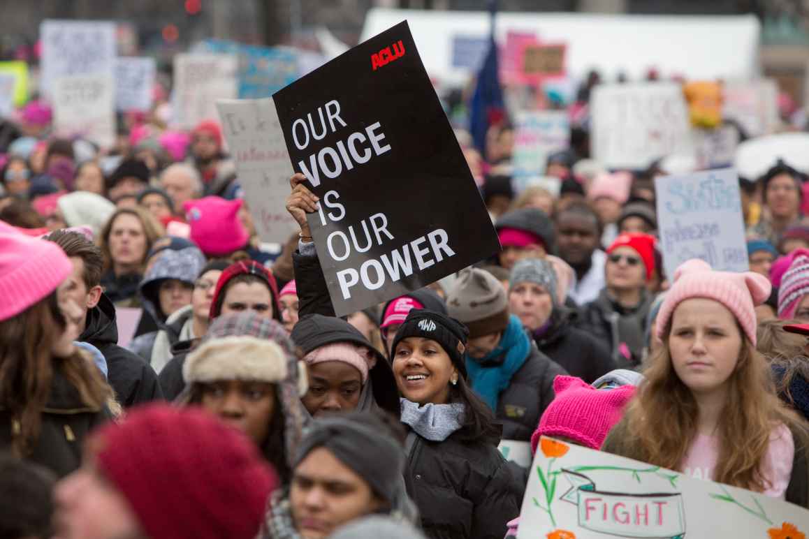 A smiling woman is holding a sign that says "Our voice is our power" in a crowded protest.