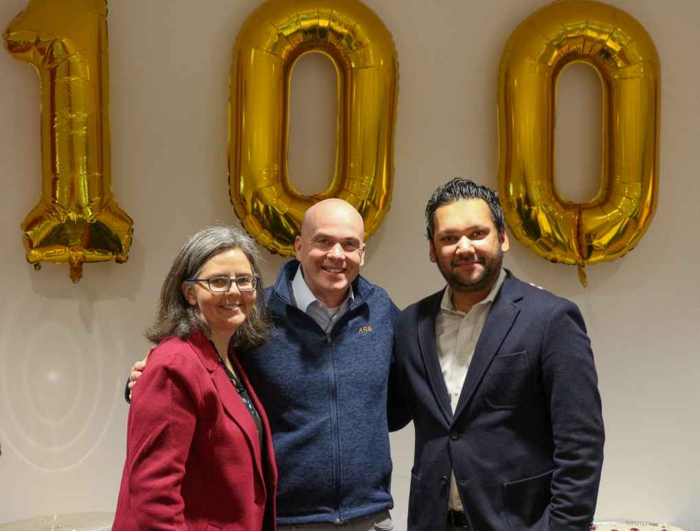 Three people standing in front of ballons of the number 100 in an office conference room